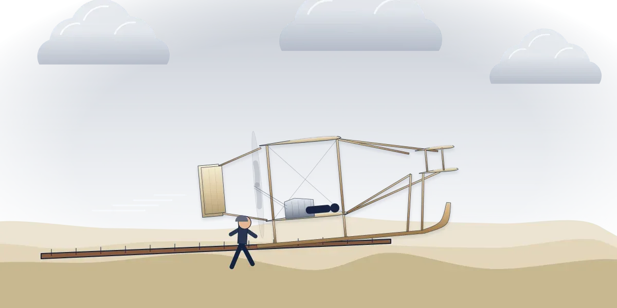 The 1903 Wright Flyer lifting off its wooden launching rail over the sand at Kitty Hawk, with a pilot lying on the lower wing and a man running alongside under a gray sky